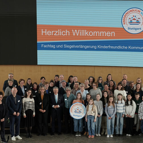 Eine Gruppe Erwachsener und Kinder steht im großen Sitzungssal im Rathaus, darunter OB Dr. Frank Nopper und die Kinderbeauftragte Maria Haller-Kindler. Kinder halten eine Attrappe des Siegels und des Stuttgarter Stadtschilds.