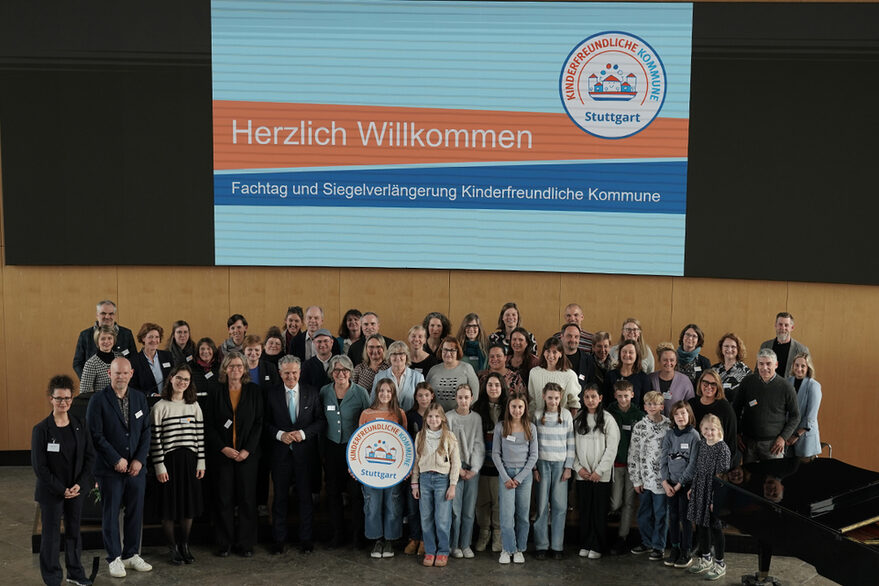 Eine Gruppe Erwachsener und Kinder steht im großen Sitzungssal im Rathaus, darunter OB Dr. Frank Nopper und die Kinderbeauftragte Maria Haller-Kindler. Kinder halten eine Attrappe des Siegels und des Stuttgarter Stadtschilds.