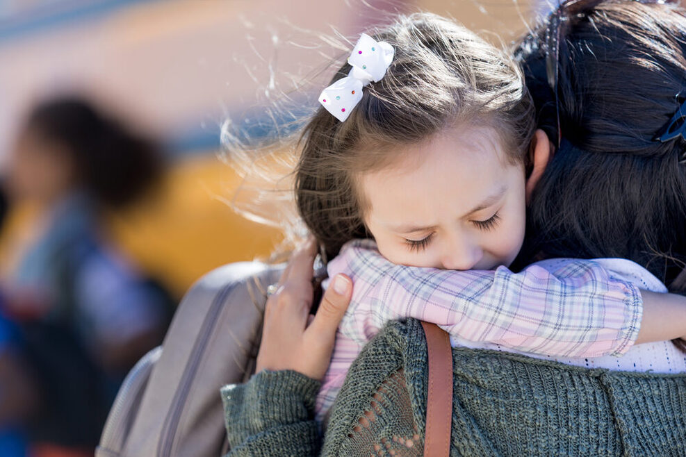 Ein kleines Mädchen mit weißer Schleife im Haar umarmt eine Frau.
