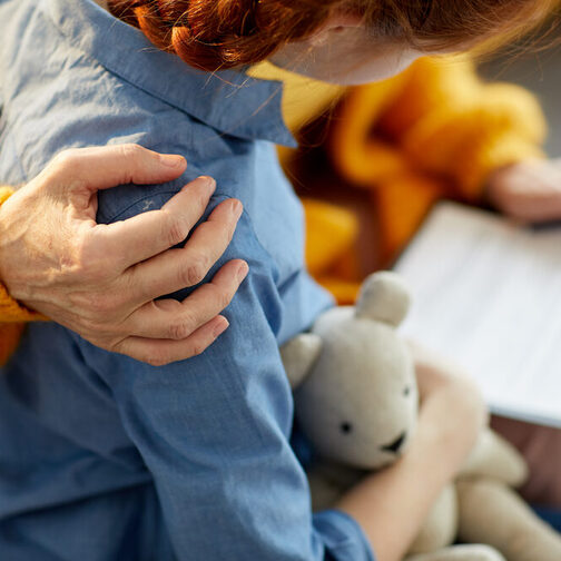 Ein Frau hält den Arm schützend um ein Mädchen. Das Kind hält einen Teddy im Arm, die Frau hält ein Formular in der Hand.