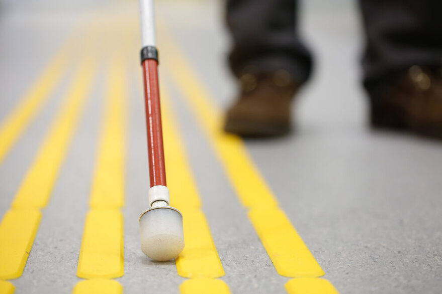 Nahaufnahme eines Blindenstocks einer blinden Person, der das Leitliniensystem auf dem Asphalt zur Orientierung nutzt