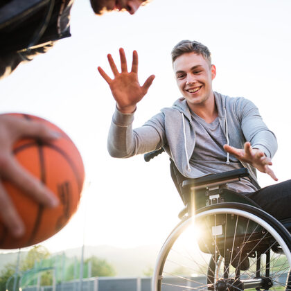 Jugendlicher im Rollstuhl spielt mit 2 Freunden draußen Basketball.