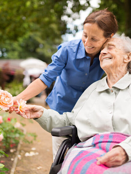 Eine ältere Dame sitzt im Rollstuhl und lächelt während eine jüngere Frau der Frau im Rollstuhl ein Rose zuneigt.