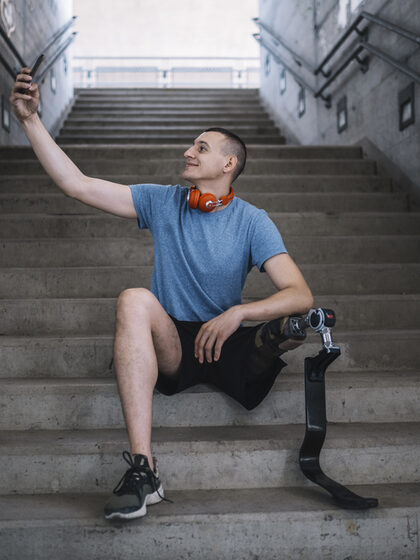 Junger Mann mit Beinprothese macht ein Selfie von sich, während er auf den Stufen eines Treppenaufgangs sitzt