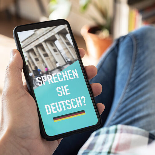 Ein Hand hält ein Smartphone. Auf Bildschirm steht "Sprechen Sie Deutsch?"