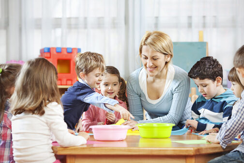 Eine Erzieherin sitzt mit Kindern an einem Tisch. Gemeinsam spielen sie ein Spiel.
