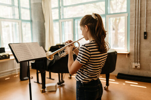 Ein Mädchen spielt Trompete. Vor ihr steht ein Notenpult. Sie trägt eine schwarze Jeans und ein  schwarzweiß gestreiftes T-Shirt.