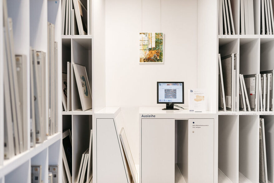 Blick auf ein Ausleiheterminal in der Stadtbibliothek am Mailänder Platz, das umrandet wird von Regalen mit Bildern der Graphotek.