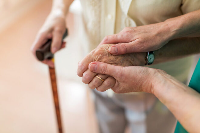 Eine jüngere Frau stützt eine ältere Frau, die einen Gehstock nutzt. Nahaufnahme der Hände.