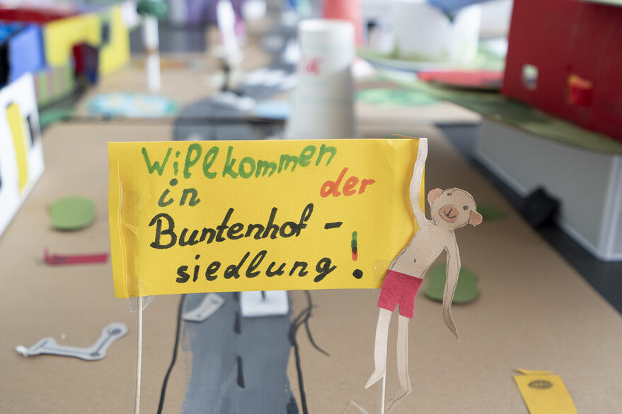 Eine gebastelte Häusersiedlung aus Pappe mit einem kleinen Plakat im Vordergrund, auf dem steht: Willkommen in der Buntenhofsiedlung.