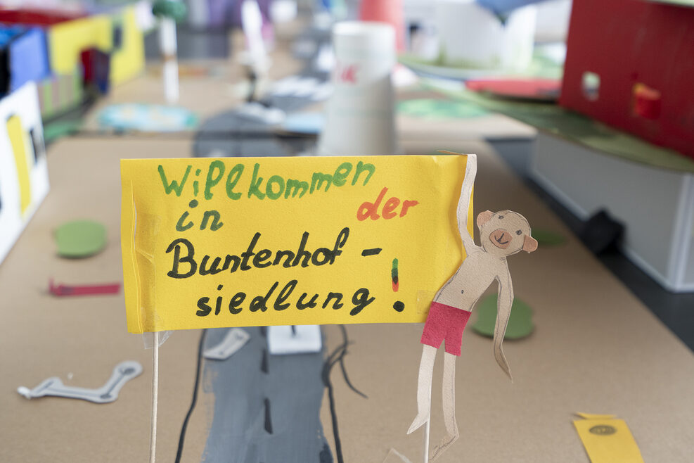 Eine gebastelte Häusersiedlung aus Pappe mit einem kleinen Plakat im Vordergrund, auf dem steht: Willkommen in der Buntenhofsiedlung.