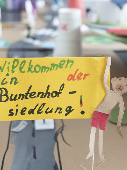 Eine gebastelte Häusersiedlung aus Pappe mit einem kleinen Plakat im Vordergrund, auf dem steht: Willkommen in der Buntenhofsiedlung.