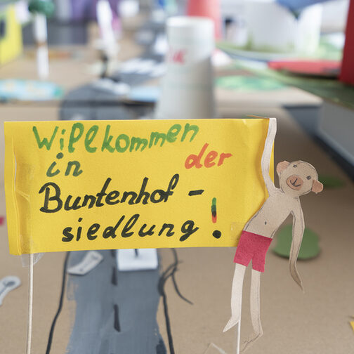 Eine gebastelte Häusersiedlung aus Pappe mit einem kleinen Plakat im Vordergrund, auf dem steht: Willkommen in der Buntenhofsiedlung.