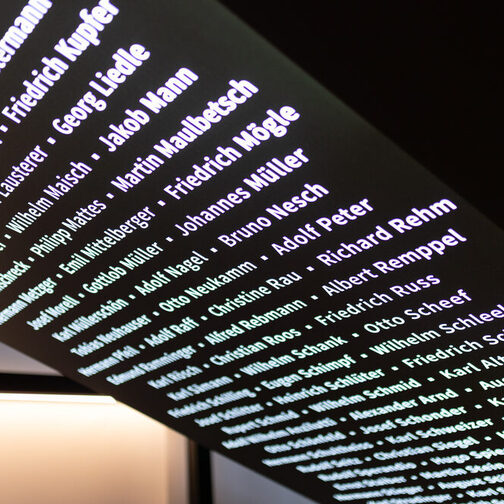 Eine schwarze, schräge Wand, an der in leuchtenden weißen Buchstaben Namen geschrieben sind.