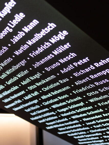 Eine schwarze, schräge Wand, an der in leuchtenden weißen Buchstaben Namen geschrieben sind.