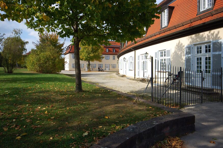 Außenansicht der Akademie Schloss Solitude. Gebäude in herbstlicher Atmosphäre mit Bäumen und Rasen.