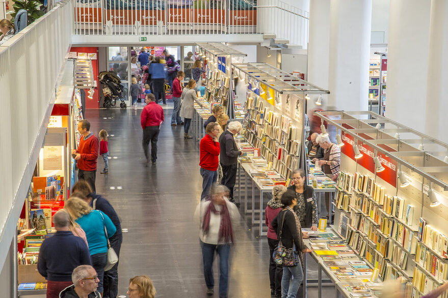 Mehrere Stände mit Büchern in einem großen Raum, Besucher schlendern hindurch.