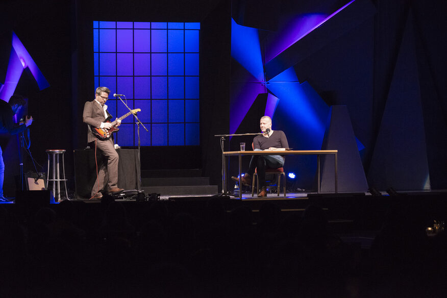 Zwei Männer auf einer Bühne: Einer sitzt an einem Tisch und liest, der andere spielt E-Gitarre.