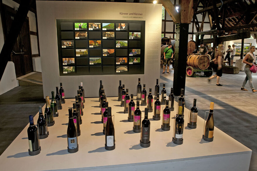 Innenansicht des Weinbaumuseums mit Fachwerkdecke. Im Vordergrund ist eine Ausstellungsfläche mit vielen Weinflaschen.