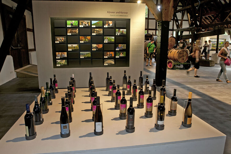 Innenansicht des Weinbaumuseums mit Fachwerkdecke. Im Vordergrund ist eine Ausstellungsfläche mit vielen Weinflaschen.