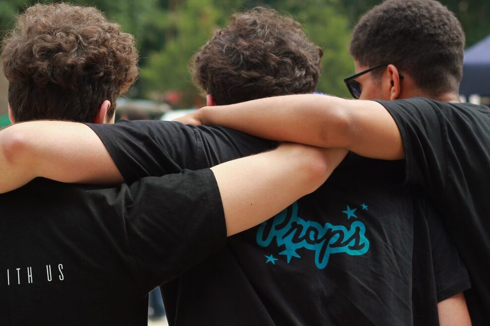 Drei junge Männer mit den Rücken zum Betrachter stehend. Auf der Rückseite der Shirts steht "Dance with us" und Props.