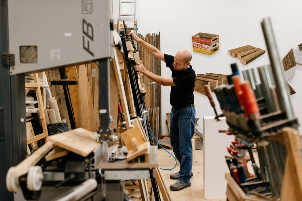 Ein Künstler steht in seinem Atelier im Kunstverein Wagenhallen, umgeben von Holzteilen, Werkzeug und angefangenen bildhauerischen Projekten.