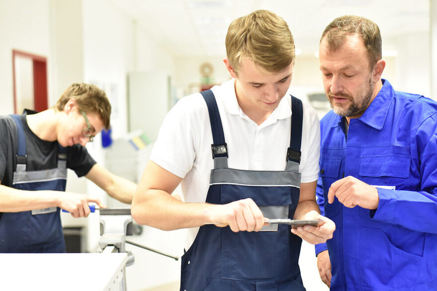 Ein älterer Mann erklärt einem jüngeren Mann etwas in einem Industriebetrieb. Im Hintergrund arbeitet ein weiterer junger Mann.