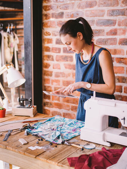 Junge Designerin steht an einer Nähmaschine und hält ein Tablet in den Händen.