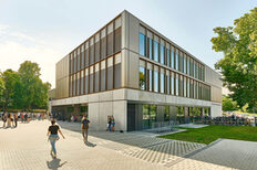 Neubau des Elly-Heuss-Knapp-Gymnasiums in Bad Cannstatt von außen mit einem massiven Sockel und bronzefarbenen Fassadenelementen aus Aluminium. Im Außenbereich vor dem Gebäude stehen Schülerinnen und Schüler.