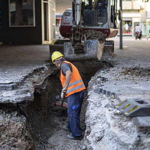 Ein Mann arbeitet mit einer Schaufel in einer Baugrube. An der Grube steht ein kleiner Bagger.