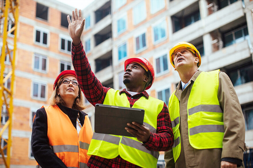 Drei Menschen, eine Frau und zwei Männer, stehen auf einer Baustelle und blicken empor, einer der Männer hat ein Tablet in der Hand.