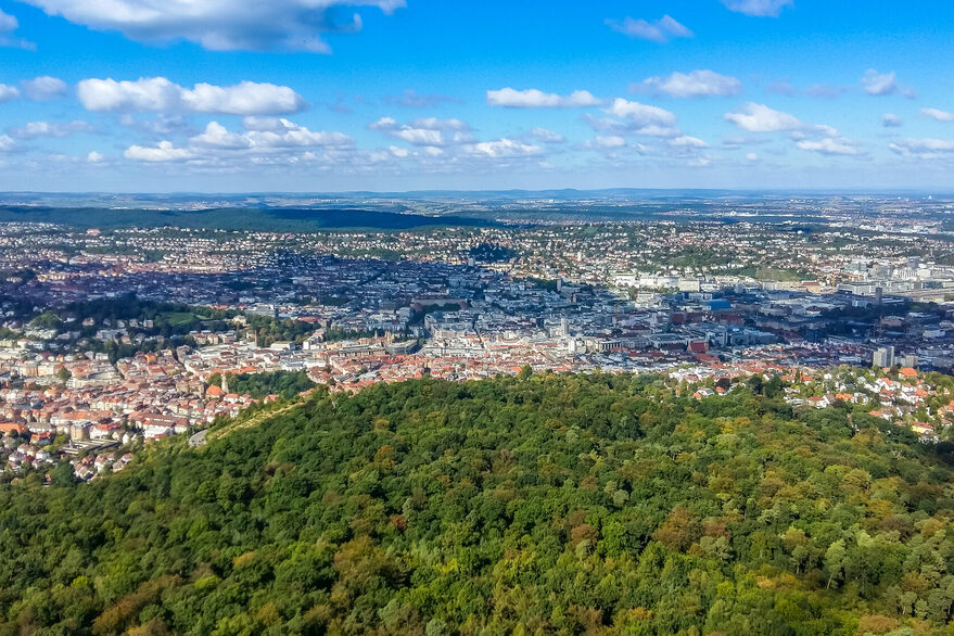 Blick über eine Stadt, im Vordergrund ist viel Wald zu sehen.