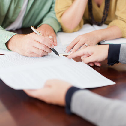 Drei Menschen sitzen an einem Schreibtisch, ein Mann unterschreibt ein Formular.