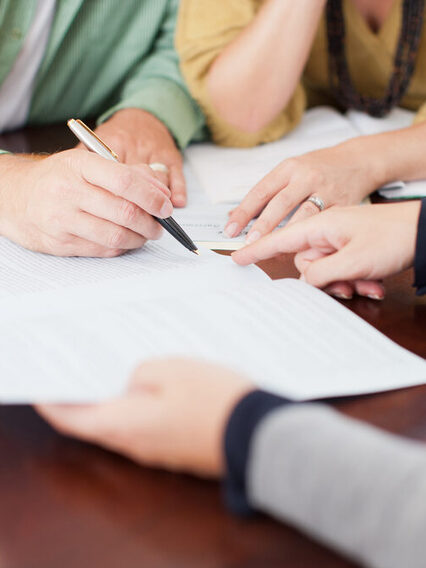 Drei Menschen sitzen an einem Schreibtisch, ein Mann unterschreibt ein Formular.