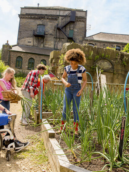 Kinder und ein Mann im Rollstuhl stehen in einem urbanen Garten mit Hochbeeten.