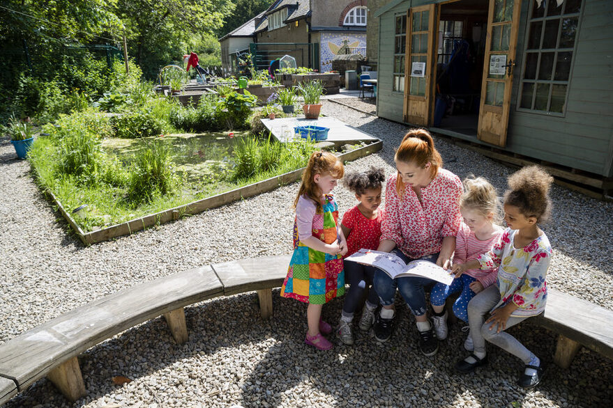 Frau sitzt mit vier Kindern auf eine halbrunden Bank in einem Hof und liest vor.