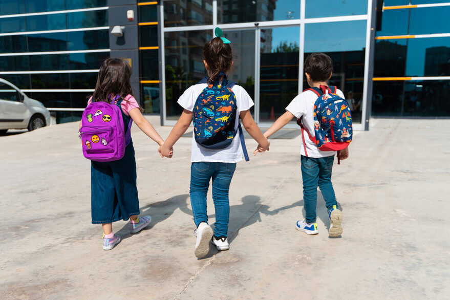 Drei Kinder mit Schulranzen halten sich an der Hand und laufen auf einen Eingang zu.