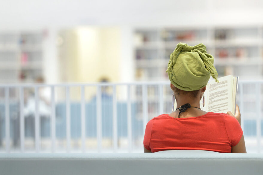 Eine Frau sitzt auf einer Bank und liest ein Buch, im Hintergrund ein heller Raum mit Bücherregalen.