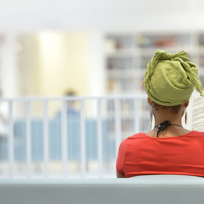 Eine Frau sitzt auf einer Bank und liest ein Buch, im Hintergrund ein heller Raum mit Bücherregalen.