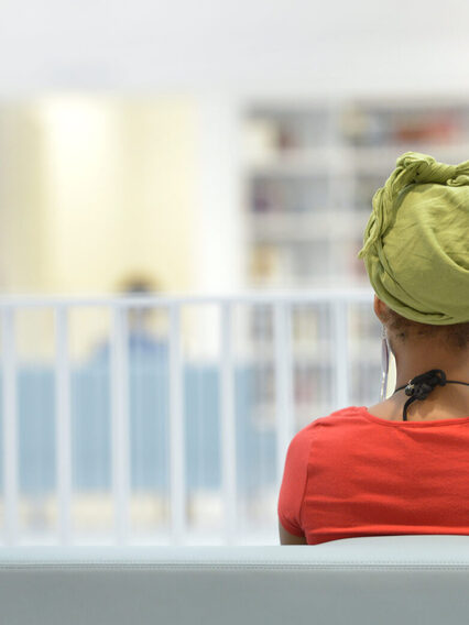 Eine Frau sitzt auf einer Bank und liest ein Buch, im Hintergrund ein heller Raum mit Bücherregalen.