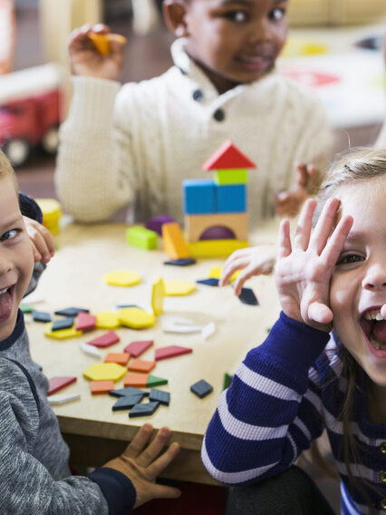 Eine Gruppe Kinder spielt an einem Tisch in der Kita. Sie blicken lachend in die Kamera.