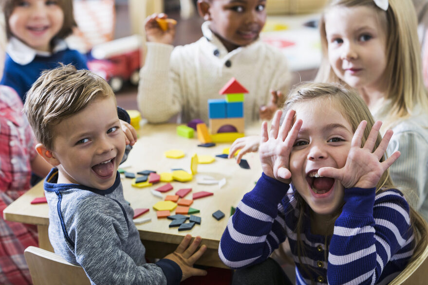 Eine Gruppe Kinder spielt an einem Tisch in der Kita. Sie blicken lachend in die Kamera.