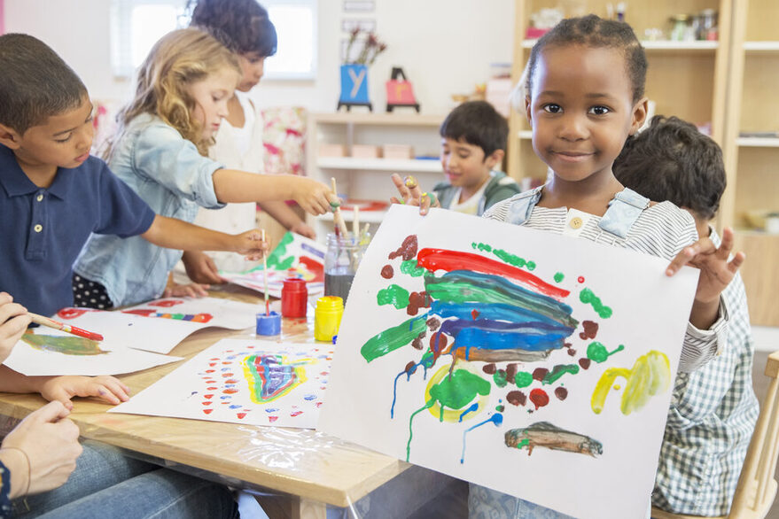 Ein Kind hält ein selbstgemaltes Bild vor sich während im Hintergrund weitere Kinder mit Wasserfarben malen.