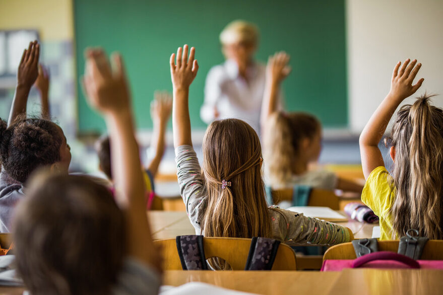 Rückansicht einer Schulklasse, die Schüler melden sich. Im Hintergrund steht eine Lehrerin vor einer Tafel.