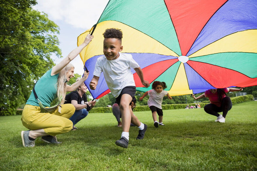 Einige Kinder spielen unter einer bunten Plane, die von Betreuerinnen gehalten wird.