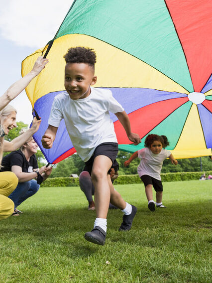 Einige Kinder spielen unter einer bunten Plane, die von Betreuerinnen gehalten wird.