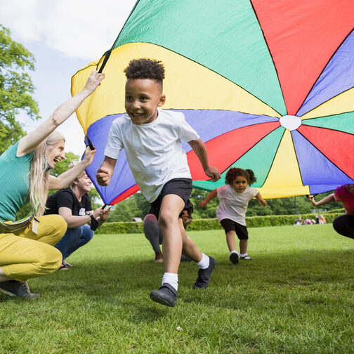Einige Kinder spielen unter einer bunten Plane, die von Betreuerinnen gehalten wird.