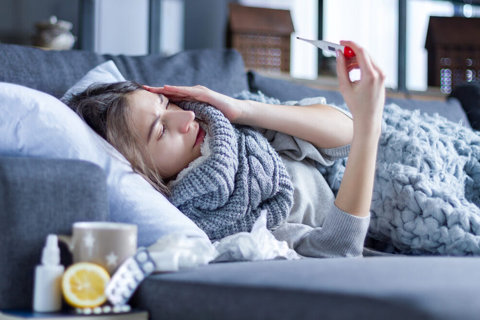 Eine Frau liegt mit einer Erkältung auf dem Sofa, eingehüllt in eine Decken, in der Hand ein Fieberthermometer