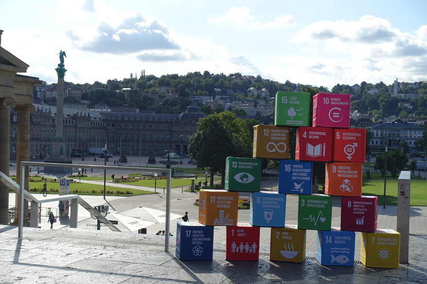 17 verschiedenfarbige Quader stehen zu einer Pyramide aufgetürmt auf der Treppe oberhalb des Schlossplatzs in Stuttgart, mit Blick in Richtung Neues Schloss. Auf den Blöcken stehen die 17 Ziele globaler Nachhaltigkeit der Vereinten Nationen.