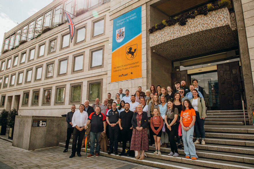 Eine Delegation von beiden Städten steht auf der Rathaustreppe des Stuttgarter Rathauses. Zu sehen sind zudem das Rathausbanner zum 75-jährigen Städtepartnerschaftsjubiläum und die britische Flagge.